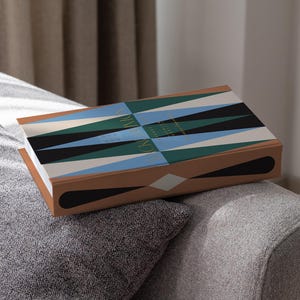 May include: A backgammon game set with a geometric design in black, blue, and green on the lid. The box has a brown border with a white diamond shape. The game is resting on a gray sofa.