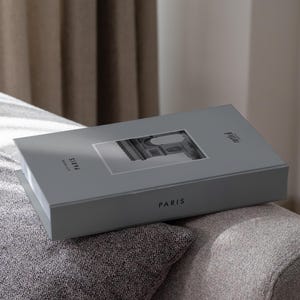 Può includere: Una scatola grigia con la scritta "PARIS" stampata sopra, con un'immagine in bianco e nero di un monumento parigino. La scatola è appoggiata su una superficie imbottita grigia.