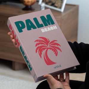 May include: A book titled "PALM BEACH" with a red palm tree illustration. The book's cover is a muted pink with green and white text. The word "artful" is printed at the bottom of the cover.
