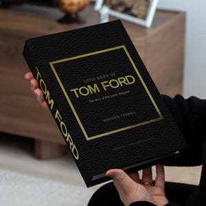 Puede incluir: Un libro negro titulado "Little Book of Tom Ford" con letras doradas y un marco dorado. La portada del libro tiene una apariencia texturizada. El libro se sostiene frente a un fondo borroso.