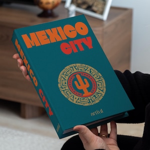 Op de afbeelding: Een turquoise boek met de titel "MEXICO CITY" in oranje en rode letters. De omslag heeft een goud en rood cirkelvormig ontwerp met een cactus. Het boek wordt in iemands handen gehouden.