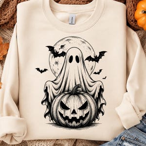 May include: A cream-colored crewneck sweatshirt featuring a black ink-style graphic of a ghost draped over a carved jack-o'-lantern pumpkin. The design includes a large full moon in the background and several flying bats surrounding the central figures.