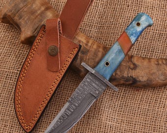 May include: A Damascus steel knife with a blue and orange handle and a brown leather sheath. The knife has a full tang construction and a decorative blade pattern. The sheath has a button closure and stitched edges. The number "703" is visible.