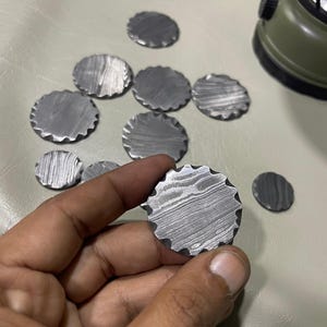 Peut inclure: Plusieurs petits objets métalliques ronds avec une surface texturée et à motifs et un bord festonné. L'un est tenu dans une main. Un objet vert est en arrière-plan.