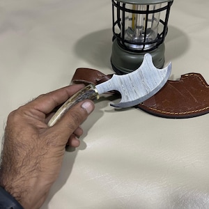 May include: A small, decorative axe with a patterned steel blade and a handle made of antler. The axe is resting on a brown leather sheath. A small, green lantern is in the background.