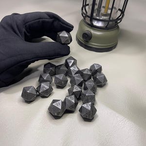 May include: A collection of dark grey, multi-sided dice with white numbering, held by a gloved hand. A green lantern with a black handle and a clear glass cover is in the background. The dice are for tabletop games.
