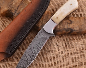 May include: A Damascus steel knife with a bone handle and a brown leather sheath. The blade has a unique swirling pattern. The handle is cream-colored with brass rivets. The sheath is brown with decorative stitching.
