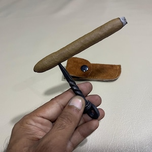 Könnte beinhalten: Eine brennende Zigarre balanciert auf einem schwarzen, gedrehten Zigarrenständer aus Metall. Im Hintergrund befindet sich ein braunes Zigarrenetui aus Leder mit einem Druckknopfverschluss. Die Zigarre ist braun mit weißer Asche.