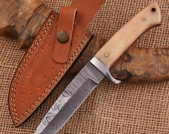 May include: A Damascus steel hunting knife with a bone handle and a brown leather sheath. The blade has a distinctive pattern, and the handle is secured with brass pins. The sheath has a belt loop and a snap closure. The knife is designed for outdoor use.