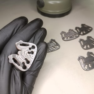Könnte beinhalten: Nahaufnahme mehrerer Metallkomponenten. Eine wird in einer behandschuhten Hand gehalten und zeigt ein einzigartiges Muster. Die anderen Komponenten sind auf einer weißen Oberfläche verstreut. Die Komponenten haben eine markante Form mit mehreren Aussparungen.