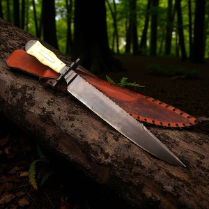 May include: A large, handmade knife with a serrated edge and a cream-colored handle, resting on a log with a brown leather sheath. The blade is long and sharp, and the knife has a black guard.