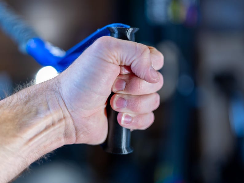 Pode incluir: Uma m&atilde;o segurando uma al&ccedil;a de exerc&iacute;cio preta e azul.
