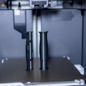 Pode incluir: Dois objetos pretos impressos em 3D, possivelmente al&ccedil;as, est&atilde;o na plataforma de uma impressora 3D.