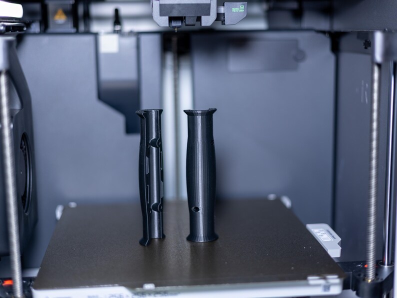 Pode incluir: Dois objetos pretos impressos em 3D, possivelmente al&ccedil;as, est&atilde;o na plataforma de uma impressora 3D.