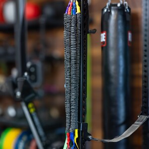 Pode incluir: Um conjunto de faixas de resist&ecirc;ncia pretas com al&ccedil;as azuis, verdes, vermelhas e amarelas penduradas em um rack de metal preto. As faixas est&atilde;o empilhadas umas sobre as outras e s&atilde;o conectadas a um clipe de metal.
