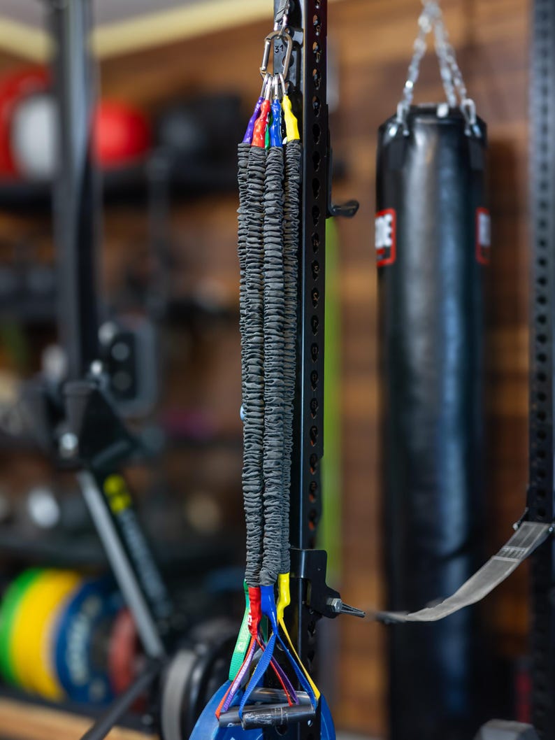 Pode incluir: Um conjunto de faixas de resist&ecirc;ncia pretas com al&ccedil;as azuis, verdes, vermelhas e amarelas penduradas em um rack de metal preto. As faixas est&atilde;o empilhadas umas sobre as outras e s&atilde;o conectadas a um clipe de metal.