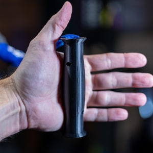 Pode incluir: Uma pega de pl&aacute;stico preto com uma superf&iacute;cie texturizada e um pequeno furo. A pega tem aproximadamente 15 cm de comprimento.