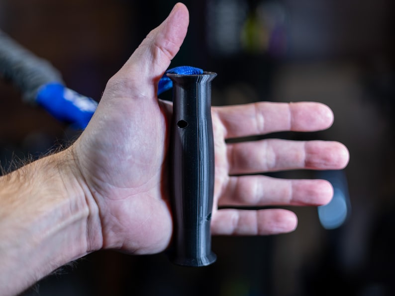 Pode incluir: Uma pega de pl&aacute;stico preto com uma superf&iacute;cie texturizada e um pequeno furo. A pega tem aproximadamente 15 cm de comprimento.