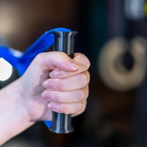 Pode incluir: Uma m&atilde;o segurando uma al&ccedil;a de exerc&iacute;cio azul e preta. A al&ccedil;a tem uma pega texturizada e um eixo de metal preto.