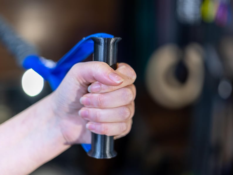 Pode incluir: Uma m&atilde;o segurando uma al&ccedil;a de exerc&iacute;cio azul e preta. A al&ccedil;a tem uma pega texturizada e um eixo de metal preto.