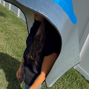 May include: A sunshade hat with a blue top and a silver reflective underside. The hat is worn by a person standing on grass. The hat is designed to provide shade from the sun.