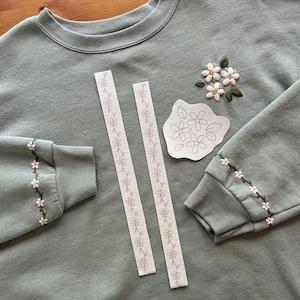 May include: Sage green sweatshirt with embroidered floral details on the sleeves and chest. Two white floral patterned ribbons and a floral pattern template are also present. The sweatshirt has a crew neck and ribbed cuffs.