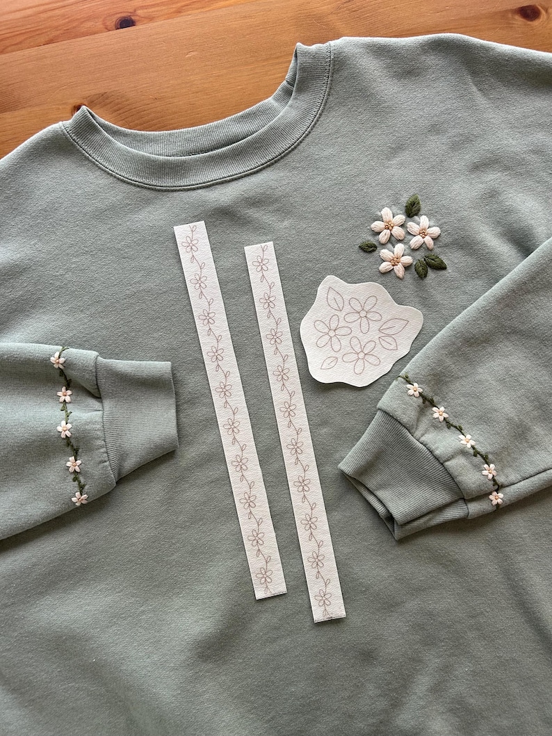 May include: Sage green sweatshirt with embroidered floral details on the sleeves and chest. Two white floral patterned ribbons and a floral pattern template are also present. The sweatshirt has a crew neck and ribbed cuffs.