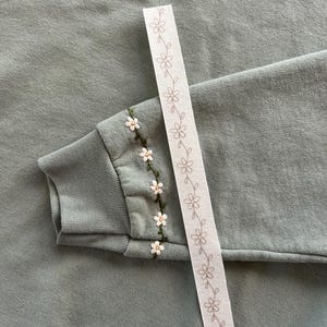 May include: Sage green sweatshirt with embroidered white and yellow flowers on the sleeve cuff. A white ribbon with a repeating floral pattern is placed diagonally across the image.