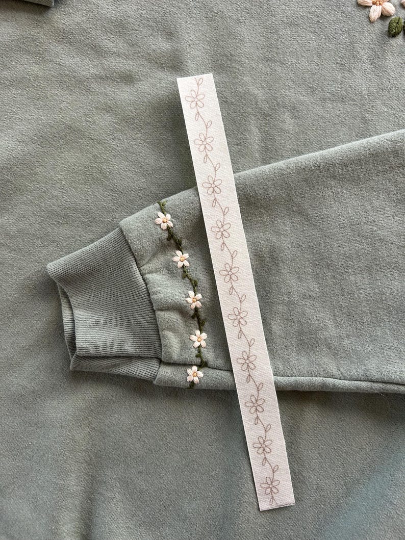 May include: Sage green sweatshirt with embroidered white and yellow flowers on the sleeve cuff. A white ribbon with a repeating floral pattern is placed diagonally across the image.