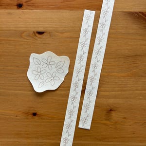 May include: A collection of floral embroidery designs on off-white fabric. The designs include a cluster of flowers and two strips with a repeating floral pattern. The designs are displayed on a wooden surface.