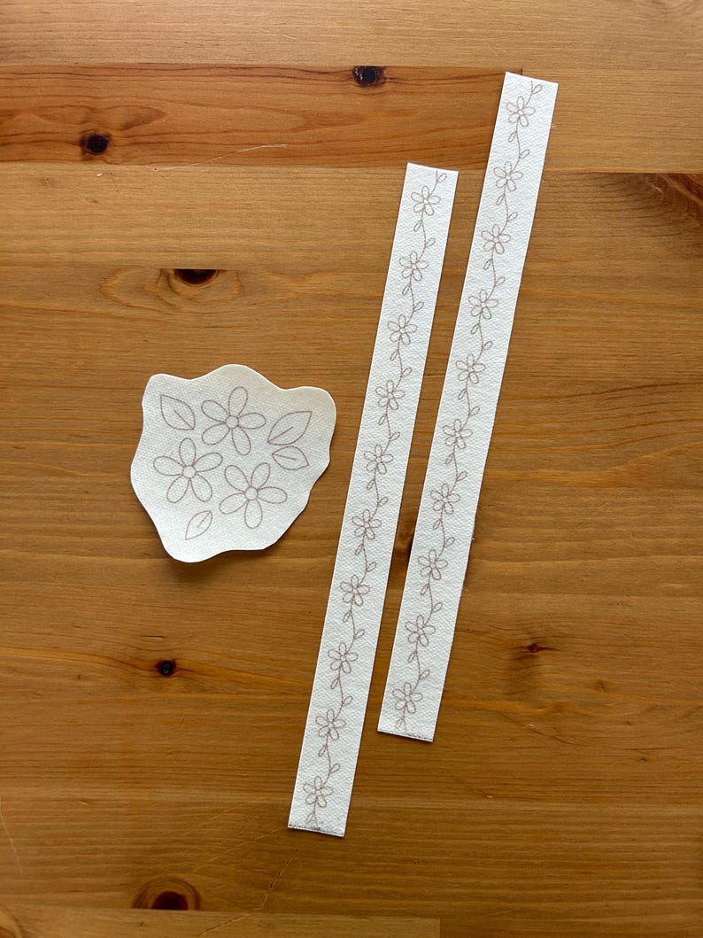 May include: A collection of floral embroidery designs on off-white fabric. The designs include a cluster of flowers and two strips with a repeating floral pattern. The designs are displayed on a wooden surface.