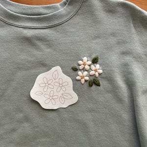 May include: Sage green sweatshirt with embroidered floral details. Features a cluster of embroidered flowers with green leaves and a separate patch with a floral outline. The sweatshirt has a crew neck.