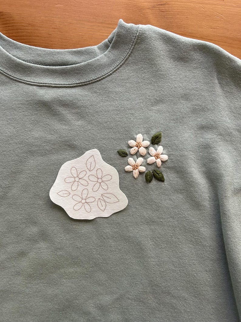 May include: Sage green sweatshirt with embroidered floral details. Features a cluster of embroidered flowers with green leaves and a separate patch with a floral outline. The sweatshirt has a crew neck.
