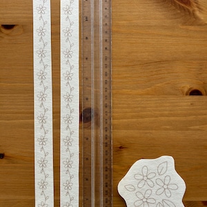 May include: Two white fabric strips with embroidered floral patterns, a clear ruler marked in inches, and a floral applique on a wooden surface. The ruler is approximately 12 inches long. The floral design is in a neutral color.