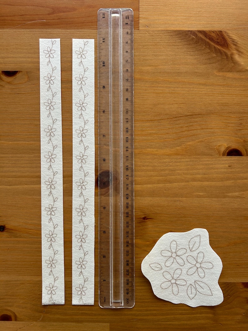 May include: Two white fabric strips with embroidered floral patterns, a clear ruler marked in inches, and a floral applique on a wooden surface. The ruler is approximately 12 inches long. The floral design is in a neutral color.