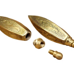 May include: Two elongated, gold-colored metal containers with pointed ends and textured, engraved surfaces. One container is open, revealing a threaded interior, while two small, matching threaded metal stoppers with rounded tops sit nearby.