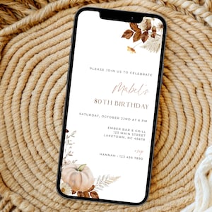 Puede incluir: Un teléfono inteligente muestra una invitación de cumpleaños digital con fondo blanco, detalles florales marrones y una ilustración de calabaza. El texto dice: Please join us to celebrate Mabel's 80th birthday, Saturday, October 22nd at 6 PM, Ember Bar & Grill, 123 Main Street, Laketown, NC 45678, RSVP Hannah 123 456 7890.