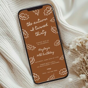Puede incluir: Un teléfono inteligente muestra una invitación digital marrón con texto blanco e ilustraciones de hojas. La invitación dice "the autumn I turned thirty" y anuncia una celebración de 30 cumpleaños con detalles del evento.