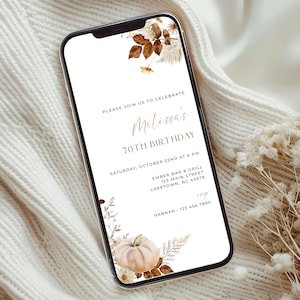 Puede incluir: Un teléfono inteligente muestra una invitación de cumpleaños. La invitación presenta el texto "Melissa's 70th Birthday" con detalles florales y de hojas en tonos marrones y beige. El teléfono está sobre un fondo blanco texturizado.