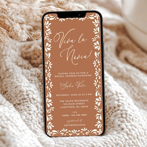May include: A smartphone displays a bridal shower invitation. The invitation features the text "Viva la Novia!" in elegant script, surrounded by a white floral border on a terracotta background. Additional details include the date, time, and location.