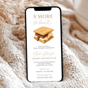 Könnte beinhalten: Ein Smartphone zeigt eine Babyparty-Einladung mit einer S'more-Grafik. Der Text lautet "S'MORE to love" und enthält Details zur Veranstaltung, wie Datum, Uhrzeit, Ort und RSVP-Informationen. Das Design ist in Brauntönen gehalten.