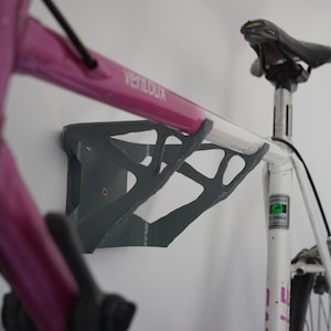 May include: A gray bike rack mounted to a white wall with a pink and white bicycle hanging on it. The bike has a black seat and a white frame with black lettering.