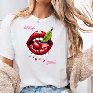 May include: White t-shirt featuring a graphic of red lips with a cherry and green leaf. The words "berry" and "good" are in pink script. The shirt is a casual, everyday wear item.