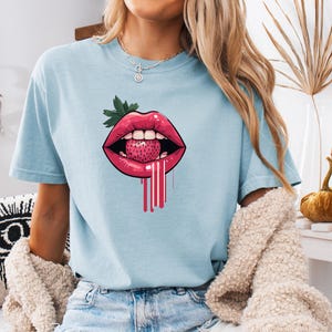 May include: Light blue t-shirt featuring a graphic of red lips with a strawberry inside. The strawberry has green leaves and pink drips. The shirt is a short-sleeved, crew neck style.