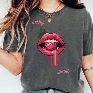 May include: Dark gray t-shirt featuring a graphic of red lips with a strawberry inside. The words "berry good" are written in pink script above and below the lips. The shirt is a casual style.
