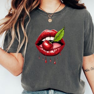 May include: Dark grey t-shirt featuring a graphic of red lips with a cherry and green leaf. The lips are dripping with red liquid. The shirt is a casual style.
