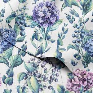 Op de afbeelding: Een stof met een bloemenpatroon met blauwe en paarse hortensia's, groene bladeren en donkerblauwe bessen op een witte achtergrond. Het ontwerp heeft een aquareleffect, met zachte schaduwen en een naturalistische stijl.