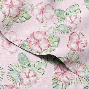May include: A light pink fabric with a repeating pattern of watercolor-style pink hibiscus flowers, green monstera leaves, and palm fronds. The floral design has yellow stamens. The fabric is folded, showing the pattern's continuity.