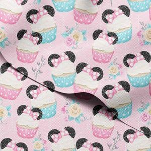 May include: Pink fabric with a repeating pattern of cupcakes with mouse ears and bows. The cupcakes have white frosting, pink and blue polka dot liners, and are interspersed with floral bouquets. The background is a solid pink color.