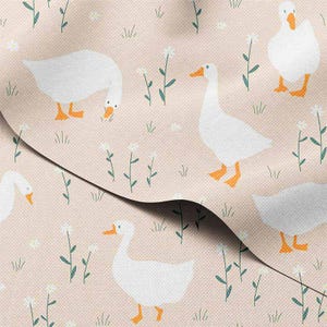 Pode incluir: Um tecido com um padrão repetido de gansos brancos com bicos e pés laranja, sobre um fundo rosa claro. O design inclui pequenas flores brancas e ilustrações de grama verde, criando um tema pastoral caprichoso.